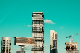 A cityscape featuring modern high-rise buildings under construction, with a prominent billboard advertisement in the foreground. A construction crane is visible, indicating urban development. The sky is a clear turquoise, adding a striking backdrop to the scene.