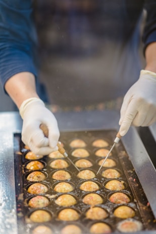 Hands wearing gloves expertly maneuver cooking tools to flip or manipulate round batter balls in a specialized pan. The cooking surface holds several semi-circular, golden-brown pastries in a rectangular grid formation. The scene suggests a food preparation or cooking process, likely in a commercial or street food setting.