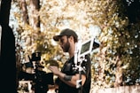 A person in a cap operating a professional video camera outdoors, surrounded by trees and foliage. The scene suggests filming or video production in a natural setting.