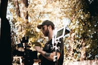 A person in a cap operating a professional video camera outdoors, surrounded by trees and foliage. The scene suggests filming or video production in a natural setting.