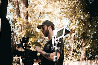 A person in a cap operating a professional video camera outdoors, surrounded by trees and foliage. The scene suggests filming or video production in a natural setting.