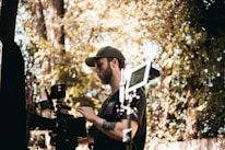 A person in a cap operating a professional video camera outdoors, surrounded by trees and foliage. The scene suggests filming or video production in a natural setting.