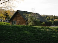 A rustic wooden cabin is nestled within lush greenery. Sunlight illuminates the leaves of a nearby tree, creating a serene and peaceful atmosphere. The cabin blends harmoniously into the natural landscape, surrounded by grass, trees, and a distant forest.