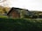 A rustic wooden cabin is nestled within lush greenery. Sunlight illuminates the leaves of a nearby tree, creating a serene and peaceful atmosphere. The cabin blends harmoniously into the natural landscape, surrounded by grass, trees, and a distant forest.