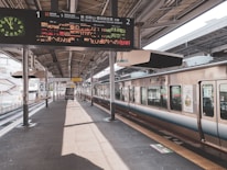 A train station platform with a train on the right side, displaying digital signboards with information in Japanese. Overhead lights and structural beams run along the platform, which is empty save for a cart near the center. A staircase and additional tracks are visible further down the platform.