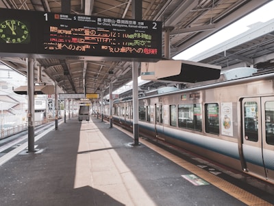 A train station platform with a train on the right side, displaying digital signboards with information in Japanese. Overhead lights and structural beams run along the platform, which is empty save for a cart near the center. A staircase and additional tracks are visible further down the platform.
