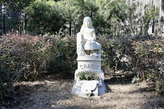 A white statue of a young girl seated upon a pedestal, surrounded by neatly trimmed bushes. The girl's features are finely detailed, and her expression appears serene. The name 'Gracie' is inscribed on the pedestal. The setting is outdoors, with rich greenery and an iron fence partially visible in the background.