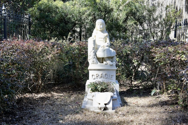 A white statue of a young girl seated upon a pedestal, surrounded by neatly trimmed bushes. The girl's features are finely detailed, and her expression appears serene. The name 'Gracie' is inscribed on the pedestal. The setting is outdoors, with rich greenery and an iron fence partially visible in the background.