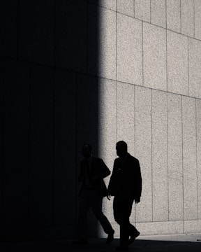 Two people in suits are walking alongside a concrete wall, with a section of the wall sharply divided between light and shadow, creating a dramatic contrast.