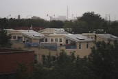 A residential area featuring modern, flat-roofed houses with large windows and solar panels. The homes are surrounded by trees and partially obscured by foliage. In the background, an urban skyline is visible under an overcast sky.