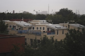 A residential area featuring modern, flat-roofed houses with large windows and solar panels. The homes are surrounded by trees and partially obscured by foliage. In the background, an urban skyline is visible under an overcast sky.