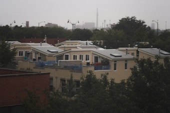 A residential area featuring modern, flat-roofed houses with large windows and solar panels. The homes are surrounded by trees and partially obscured by foliage. In the background, an urban skyline is visible under an overcast sky.