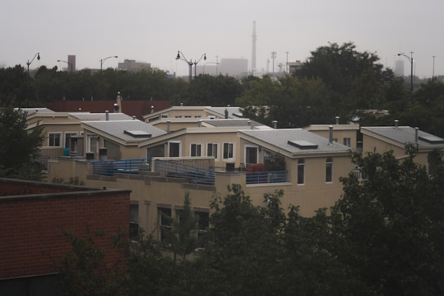 A residential area featuring modern, flat-roofed houses with large windows and solar panels. The homes are surrounded by trees and partially obscured by foliage. In the background, an urban skyline is visible under an overcast sky.