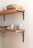 Wooden shelves mounted on a wall hold a collection of ceramic dishes and mugs. The plates and bowls are stacked neatly on the top shelf, while the bottom shelf displays several uniquely patterned mugs with a speckled design. The shelves are supported by black metal brackets, adding a touch of industrial style to the minimalist arrangement.