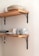 Wooden shelves mounted on a wall hold a collection of ceramic dishes and mugs. The plates and bowls are stacked neatly on the top shelf, while the bottom shelf displays several uniquely patterned mugs with a speckled design. The shelves are supported by black metal brackets, adding a touch of industrial style to the minimalist arrangement.