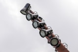 Several large spotlights are mounted on a pole against a cloudy sky. The lights are cylindrical and appear to be made of metal with an industrial look.