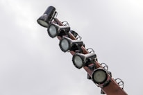 Several large spotlights are mounted on a pole against a cloudy sky. The lights are cylindrical and appear to be made of metal with an industrial look.