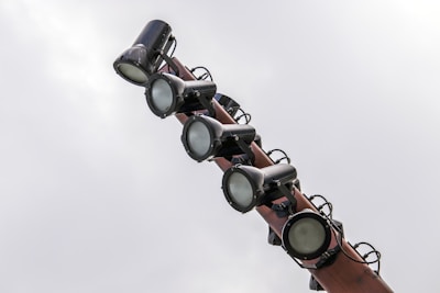 Several large spotlights are mounted on a pole against a cloudy sky. The lights are cylindrical and appear to be made of metal with an industrial look.