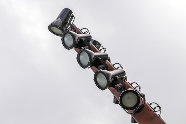 Several large spotlights are mounted on a pole against a cloudy sky. The lights are cylindrical and appear to be made of metal with an industrial look.