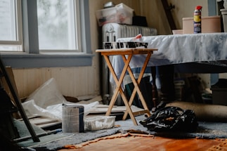 A cluttered room with painting supplies scattered around, including a can of paint, a roller tray with paint, a folding table, and a spray can. Natural light filters through a window, partially illuminating the space. The floor is covered with a worn carpet, and the background shows items like a radiator and various boxes.