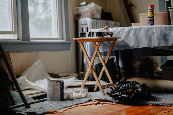 A cluttered room with painting supplies scattered around, including a can of paint, a roller tray with paint, a folding table, and a spray can. Natural light filters through a window, partially illuminating the space. The floor is covered with a worn carpet, and the background shows items like a radiator and various boxes.