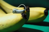 A close-up view of bananas with a hearing aid resting on top. The bananas are ripe with some brown spots on the peel, and the hearing aid is silver and slightly reflective.
