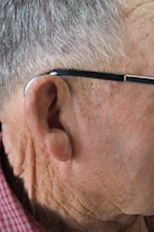 Close-up of the right side of an elderly man's head, focusing on his ear, gray hair, a hearing aid, and eyeglasses with a black frame. He is wearing a red checkered shirt.