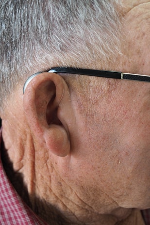 Close-up of the right side of an elderly man's head, focusing on his ear, gray hair, a hearing aid, and eyeglasses with a black frame. He is wearing a red checkered shirt.