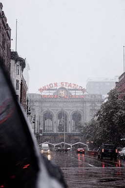 A historic train station with a prominent sign reading 'Union Station' and 'Travel by Train' in glowing red letters. The architecture is grand, featuring large arched windows and a classic clock. The street in front is wet, suggesting recent rain, and several cars with headlights on are visible, indicating low-light or overcast conditions. The scene has an urban feel with a mix of buildings and a hint of greenery along the side.