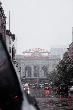 A historic train station with a prominent sign reading 'Union Station' and 'Travel by Train' in glowing red letters. The architecture is grand, featuring large arched windows and a classic clock. The street in front is wet, suggesting recent rain, and several cars with headlights on are visible, indicating low-light or overcast conditions. The scene has an urban feel with a mix of buildings and a hint of greenery along the side.