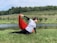 A person is performing a dance or yoga pose on a grassy field near a calm body of water. They hold a flowing red fabric in one hand, which adds a sense of movement to the scene. The background features rolling hills covered with lush green trees and a clear blue sky.