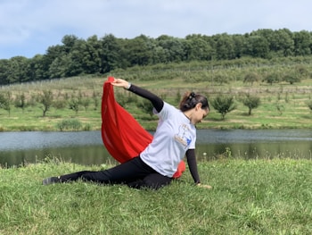 A person is performing a dance or yoga pose on a grassy field near a calm body of water. They hold a flowing red fabric in one hand, which adds a sense of movement to the scene. The background features rolling hills covered with lush green trees and a clear blue sky.