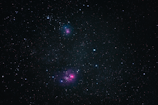A vast expanse of outer space filled with countless stars, including two noticeable clusters of pinkish nebulae. The dark background provides a stark contrast to the vibrant colors of the nebulae and the scattered azure and white stars.