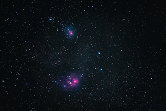A vast expanse of outer space filled with countless stars, including two noticeable clusters of pinkish nebulae. The dark background provides a stark contrast to the vibrant colors of the nebulae and the scattered azure and white stars.