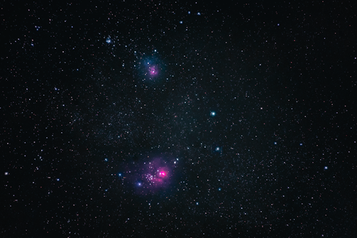 A vast expanse of outer space filled with countless stars, including two noticeable clusters of pinkish nebulae. The dark background provides a stark contrast to the vibrant colors of the nebulae and the scattered azure and white stars.