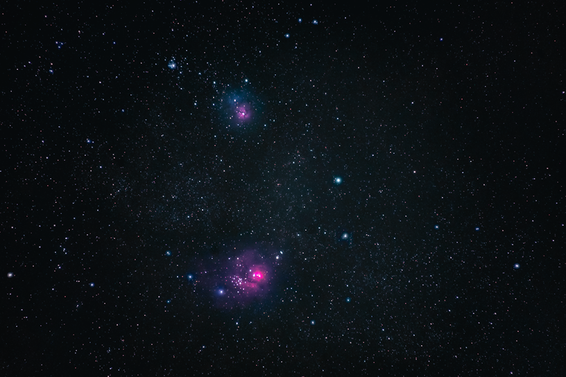 A vast expanse of outer space filled with countless stars, including two noticeable clusters of pinkish nebulae. The dark background provides a stark contrast to the vibrant colors of the nebulae and the scattered azure and white stars.