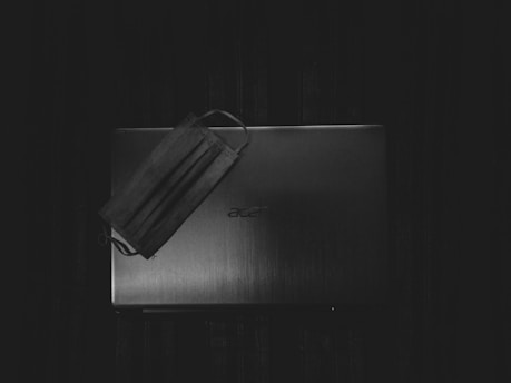 A laptop with a closed lid placed on a dark surface. A face mask is laid partially over the laptop, suggesting themes of work-from-home or remote environments.