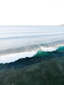 A serene view of the ocean with gentle waves rolling in. The water is a mix of deep blue and green hues, and the surface reflects the light, creating a sparkling effect. In the distance, the horizon is visible with a hint of land on the right side.