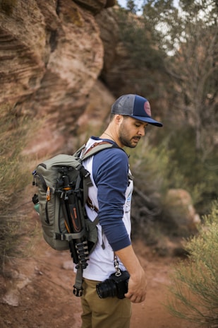 A person wearing a cap and a two-tone shirt is hiking in a natural setting. They are carrying a large backpack equipped with a camera and a tripod attached to it. The background consists of rocky terrain and some shrubs.