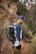 A person wearing a cap and a two-tone shirt is hiking in a natural setting. They are carrying a large backpack equipped with a camera and a tripod attached to it. The background consists of rocky terrain and some shrubs.