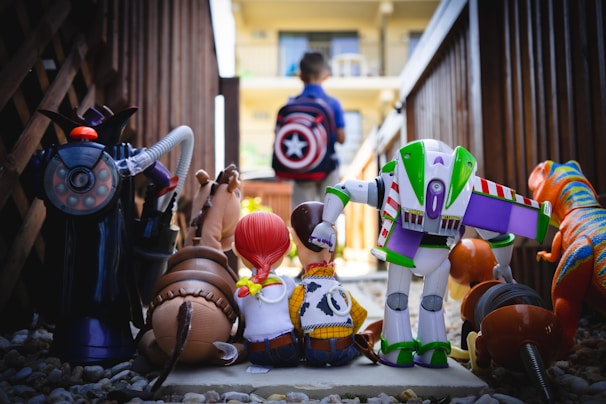 A group of toy figures, including a robot and a cowgirl, are facing away, sitting on gravel with a boy walking away in the distance wearing a backpack with a red and white star emblem. The setting seems to be an outdoor walkway framed by wooden fences.
