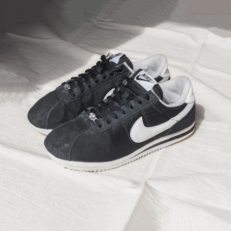 A pair of black and white Nike sneakers is positioned on a light-colored fabric surface. The shoes have a classic design with a prominent white Nike swoosh logo on the sides and black laces. They are well-lit, casting soft shadows on the textured background.
