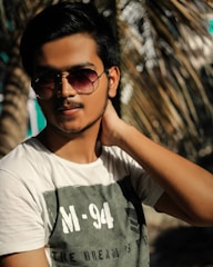A person with short black hair wearing sunglasses and a white T-shirt with the text 'M-94 The Dream Is'. The background features blurred palm leaves, suggesting a warm and sunny setting.
