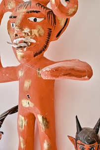 A papier-mâché figure with a menacing expression, painted primarily in red with black accents and gold patches. The figure has exaggerated features including large horns, prominent facial hair details, and a protruding tongue. Below the larger figure, a smaller, similarly styled figure with dark horns and a menacing look is visible.