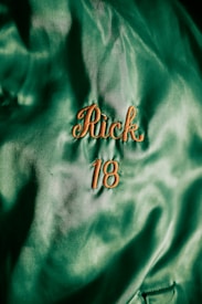 A close-up shot of a green fabric with the name 'Rick' and the number '18' embroidered in orange thread. The fabric appears textured and has a slightly shiny surface, suggesting it might be satin or similar material.
