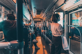 Passengers are seated and standing inside a dimly lit bus. The focus is on the interior of the bus, with handrails and seating visible. A person is standing near the door on the right, holding a bag. Advertisements and information displays are above the windows.