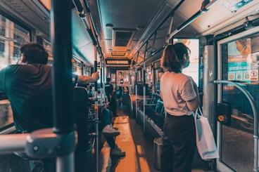 Passengers are seated and standing inside a dimly lit bus. The focus is on the interior of the bus, with handrails and seating visible. A person is standing near the door on the right, holding a bag. Advertisements and information displays are above the windows.