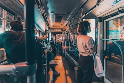 Passengers are seated and standing inside a dimly lit bus. The focus is on the interior of the bus, with handrails and seating visible. A person is standing near the door on the right, holding a bag. Advertisements and information displays are above the windows.