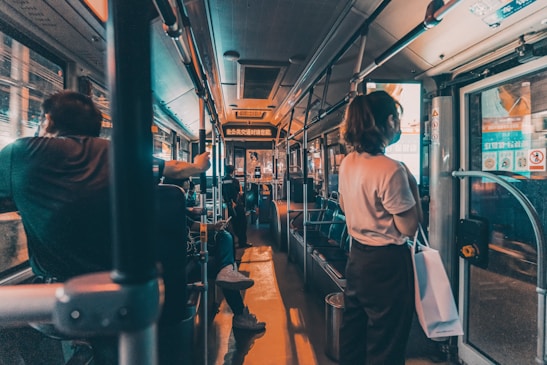 Passengers are seated and standing inside a dimly lit bus. The focus is on the interior of the bus, with handrails and seating visible. A person is standing near the door on the right, holding a bag. Advertisements and information displays are above the windows.