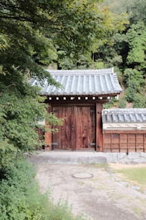 A traditional wooden gate with large doors and a tiled roof is set amidst lush greenery. The structure is flanked by wooden walls and surrounded by dense trees, creating a serene and picturesque scene.